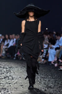 Runway model in an all-black avant-garde outfit with a giant floppy hat, long gloves, and lace-up boots, walking the catwalk.