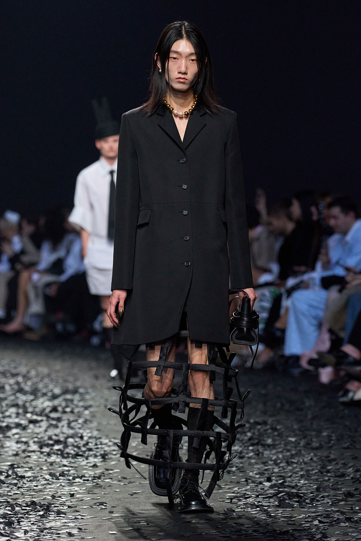 Model walking the runway in a black blazer-style coat with a button front, paired with a chunky pearl necklace and a strapped black leg harness accessory.