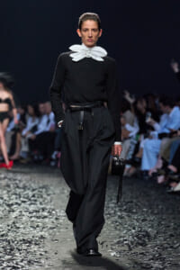 Model walks a runway in a black ensemble with a large white sculptural neck accessory, wide-leg pants, and a black belt; holds a small black fringed handbag.