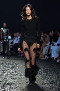 Model walks the runway in a black fitted top with a sheer, embellished skirt wearing chunky boots and a statement necklace. Concentrated audience in the background shows a fashion show setting.