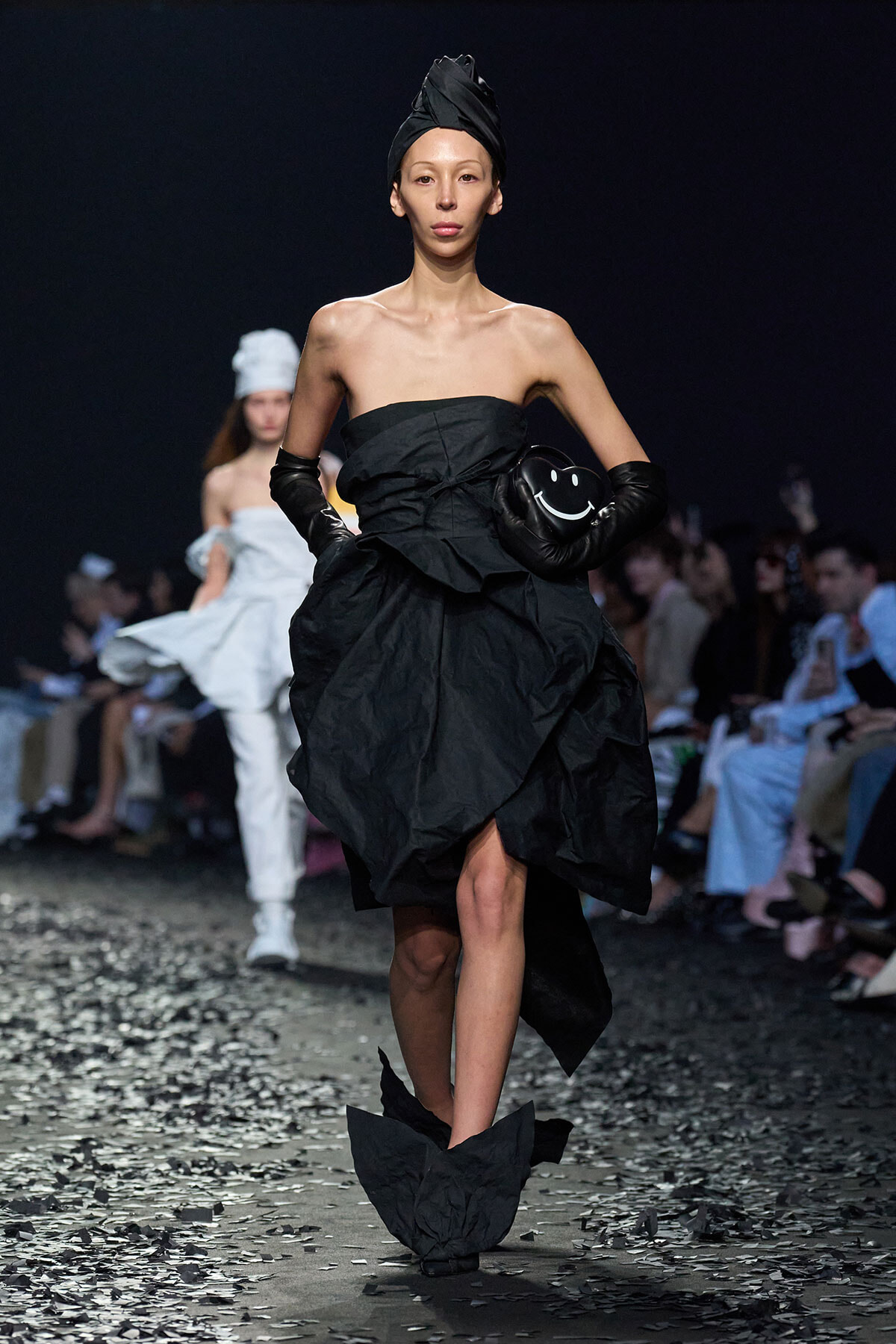 Model walks a runway in a black strapless, sculptural gown with a wrapped headpiece and long black gloves, holding a black smiley-clutch as confetti sits on the floor.