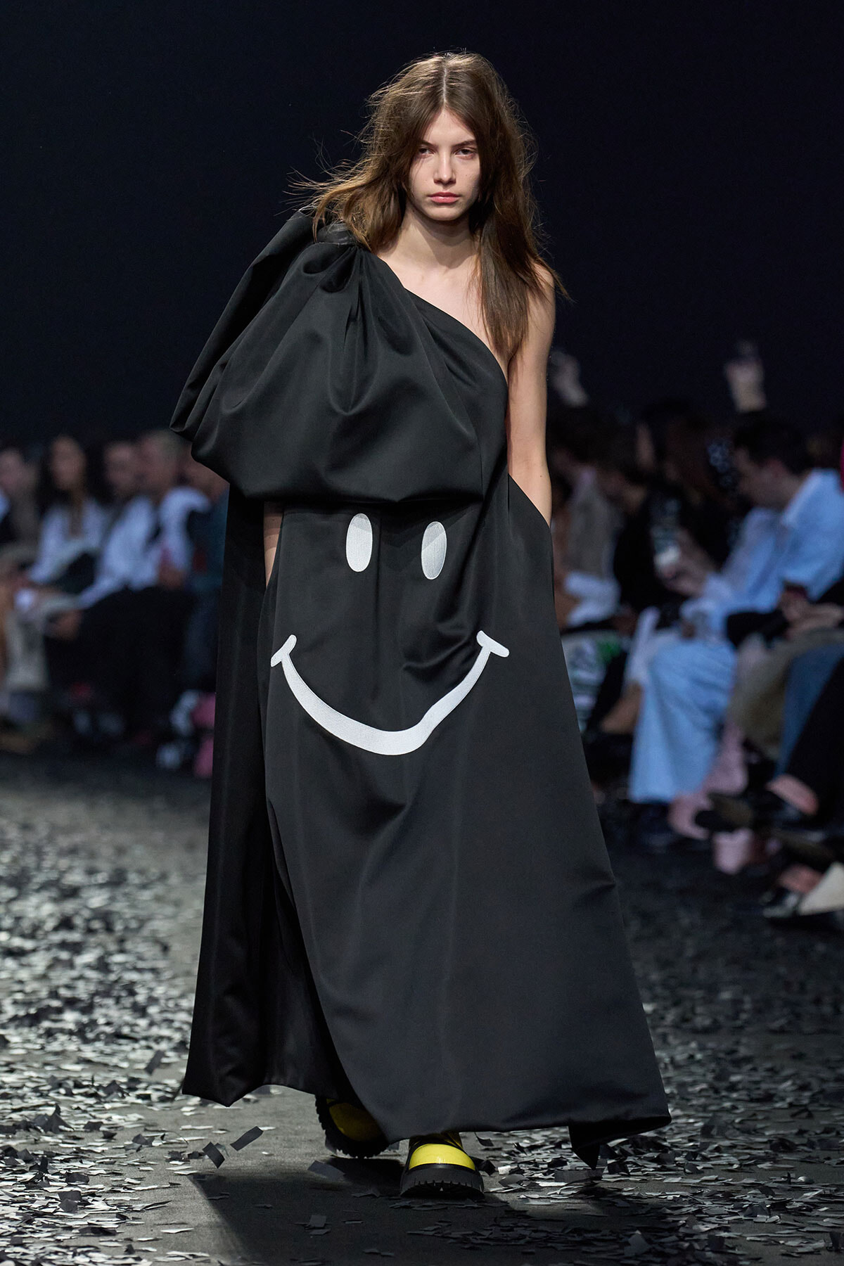 Fashion model walks a runway in a long black one-shoulder dress featuring a large white smiley face graphic, paired with bright yellow shoes; audience blurred in the background.