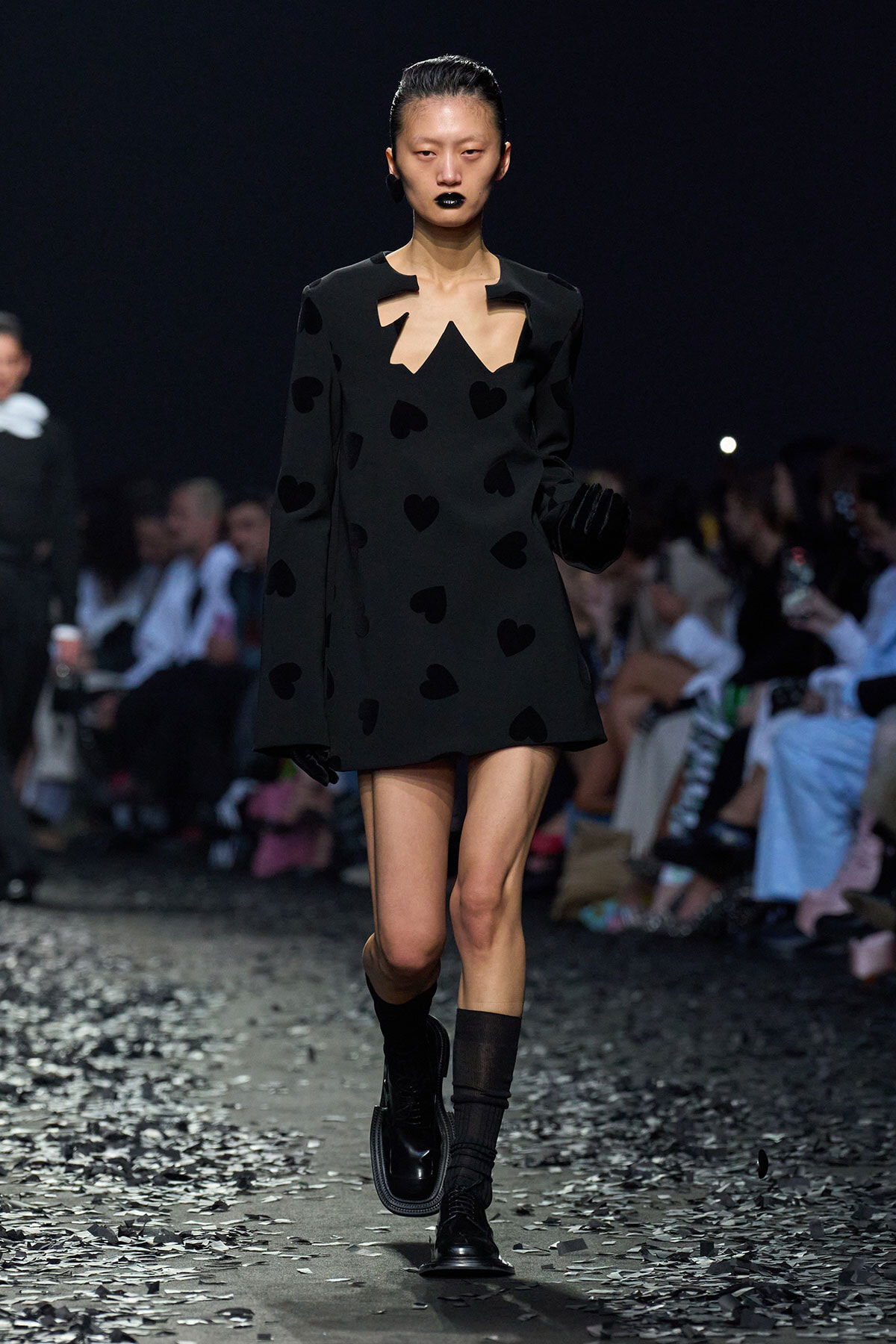 Model walking a runway in a black mini dress with heart shapes, cutout neckline, black gloves and boots.