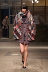 Model walking a runway in a oversized plaid coat with a large matching scarf, in burgundy, navy, and cream tones; black flats.
