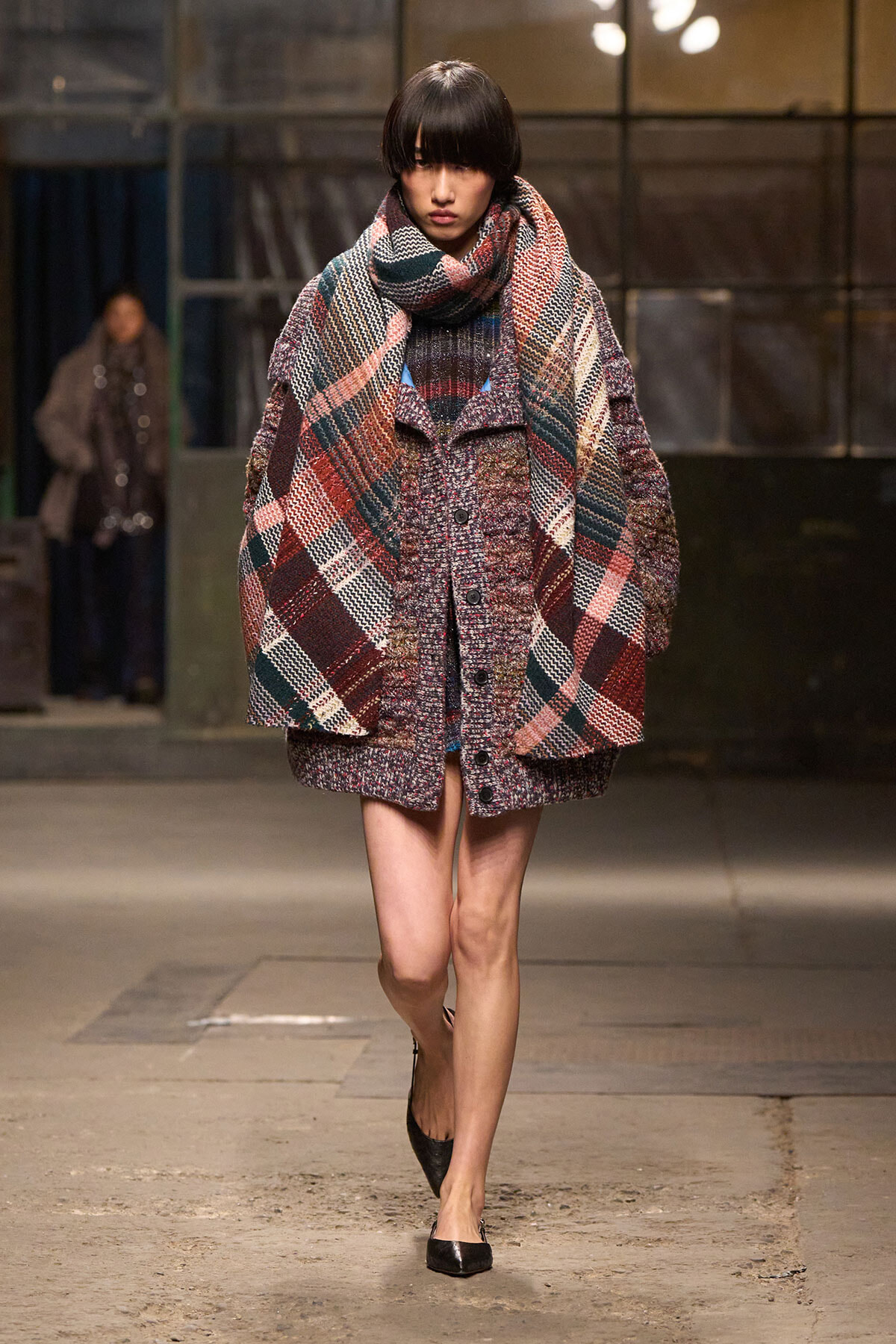Model walking a runway in a oversized plaid coat with a large matching scarf, in burgundy, navy, and cream tones; black flats.