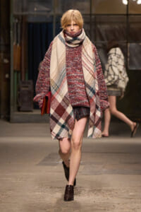 Model walks a runway in a large plaid wool coat with a matching scarf over a burgundy knit sweater and short skirt, brown boots.