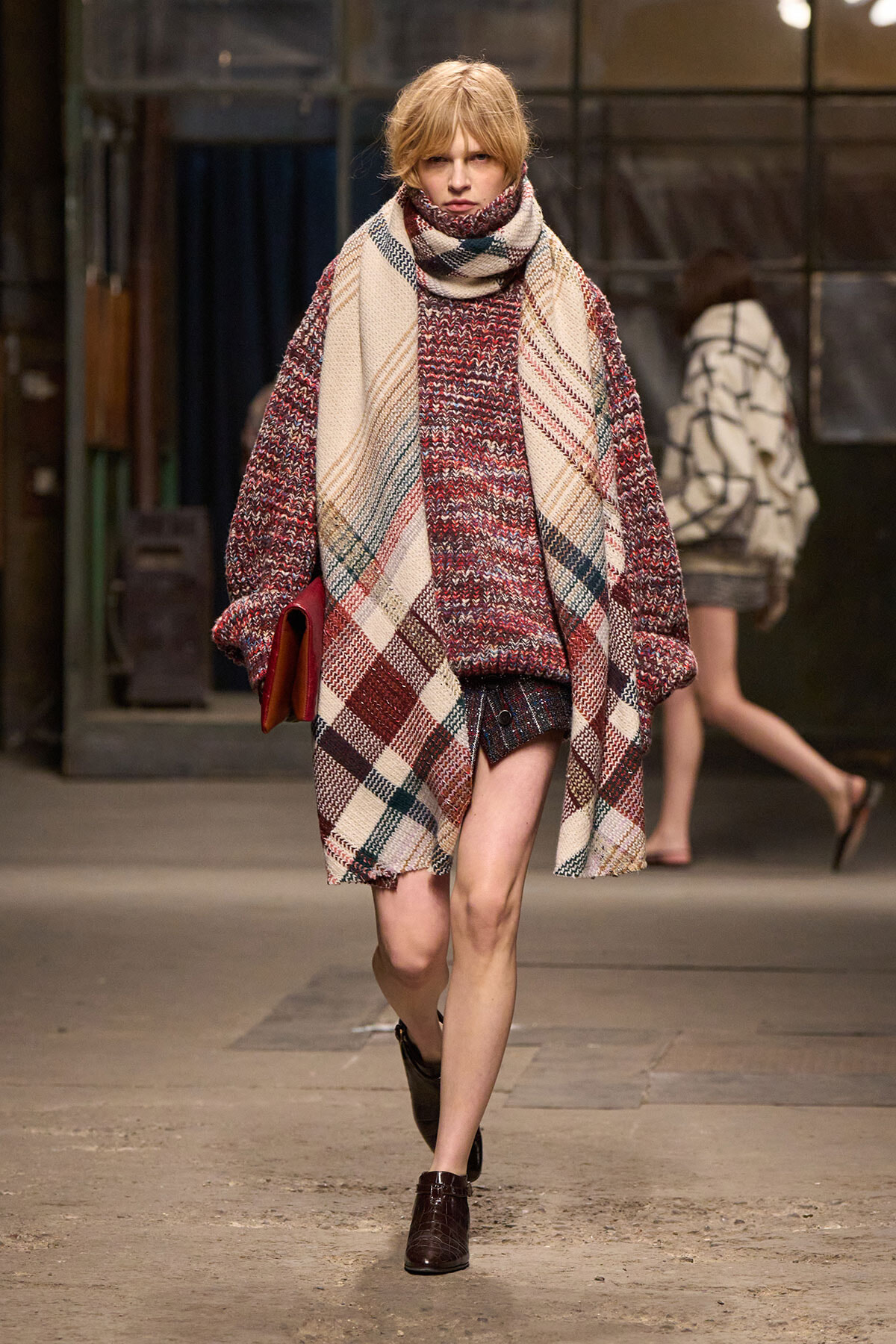 Model walks a runway in a large plaid wool coat with a matching scarf over a burgundy knit sweater and short skirt, brown boots.
