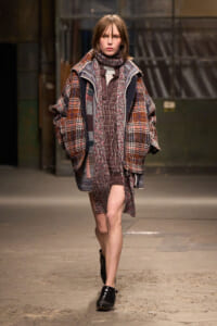 Model on a runway wearing a chunky plaid coat with a matching knit scarf and layered textures in earthy reds and browns.