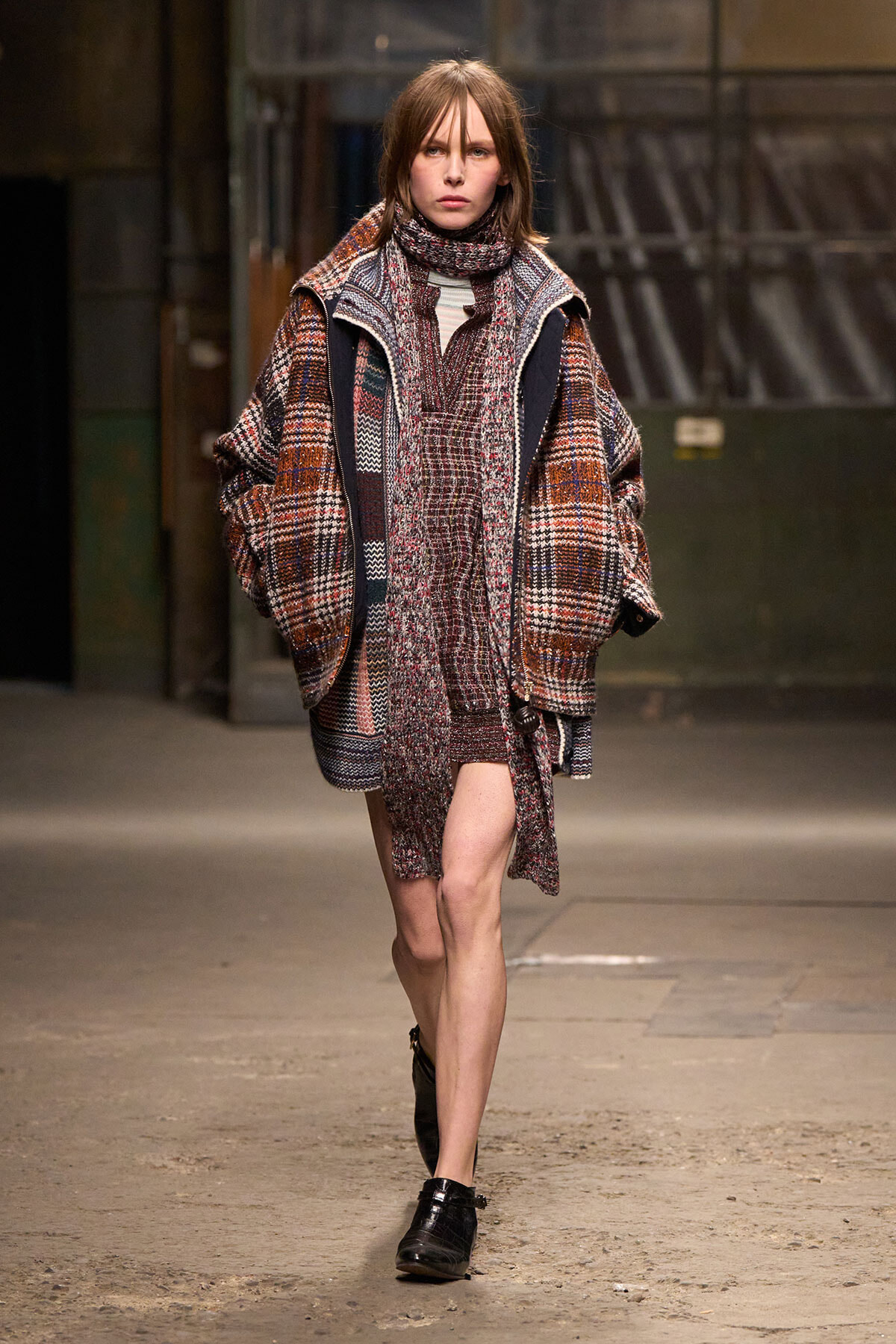 Model on a runway wearing a chunky plaid coat with a matching knit scarf and layered textures in earthy reds and browns.