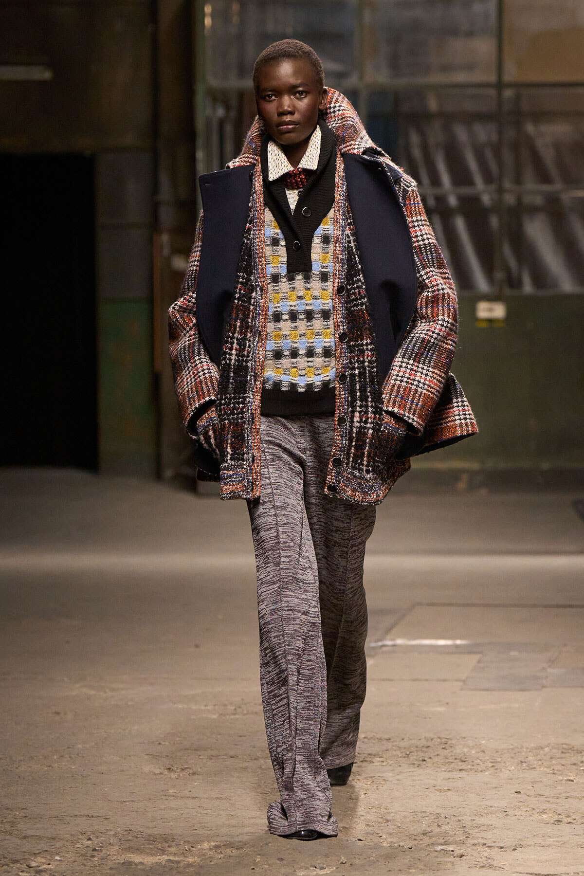Person walking on a runway in an oversized multicolored plaid coat over a checkered knit vest and wide gray trousers.