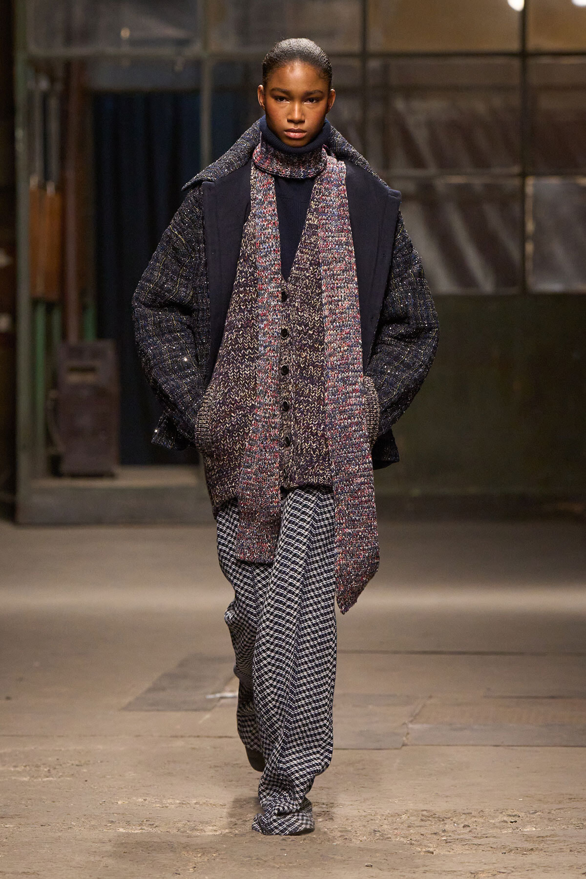 Model walking a runway in an oversized textured coat with a chunky knit scarf and wide-leg checkered pants.