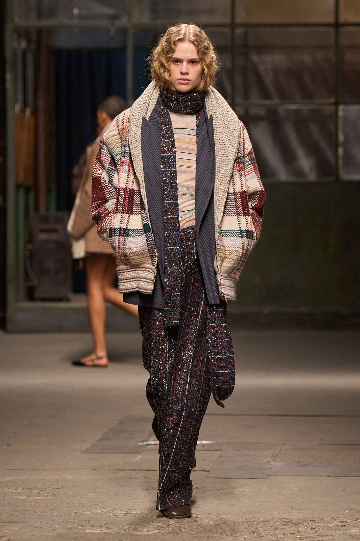 Model walks a fashion runway in layered winter-coat ensemble with a chunky plaid cardigan, scarf, and sparkling patterned trousers.