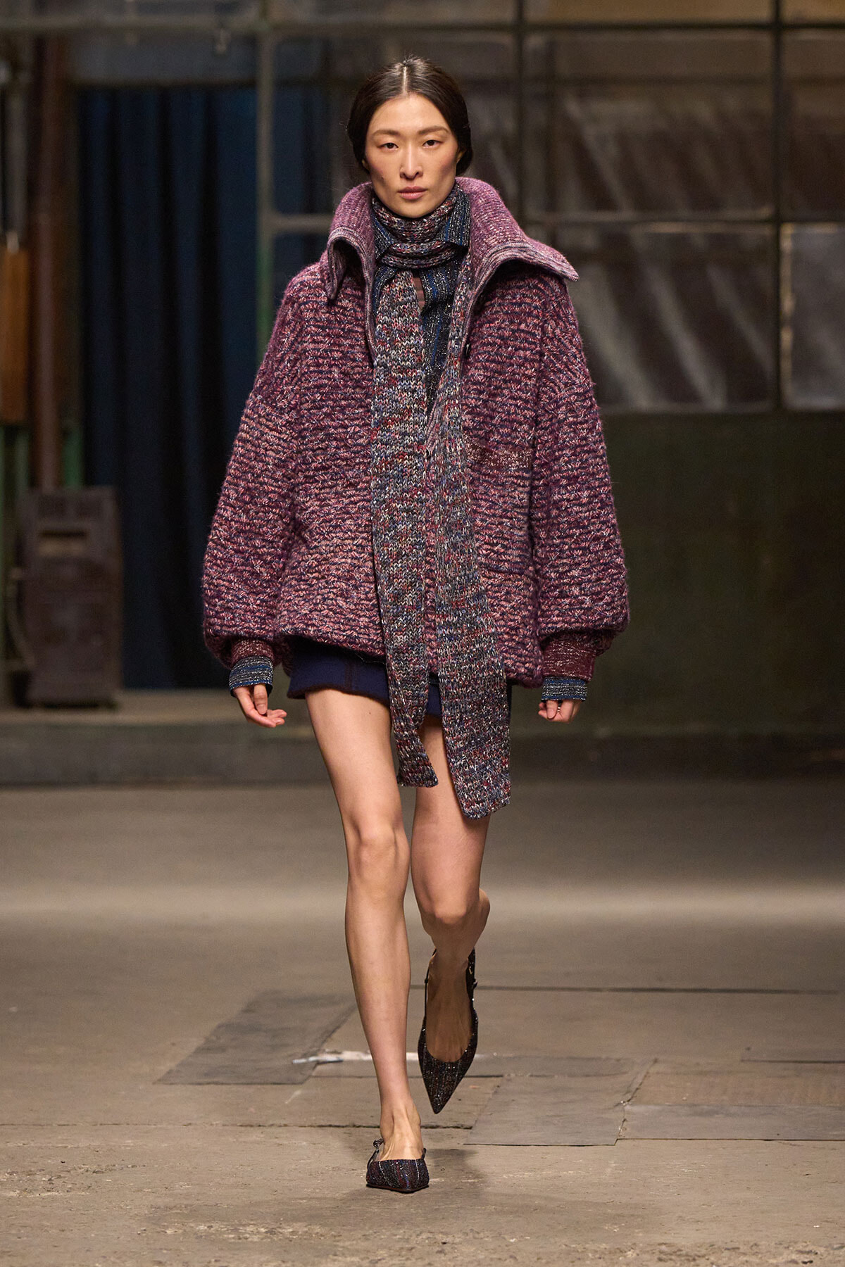 Model walks a fashion runway in an oversized maroon knit coat with matching scarf and pointed heels.