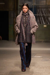 Model walking a runway in an oversized textured coat over a plaid blazer and scarf, with patterned wide-leg pants.