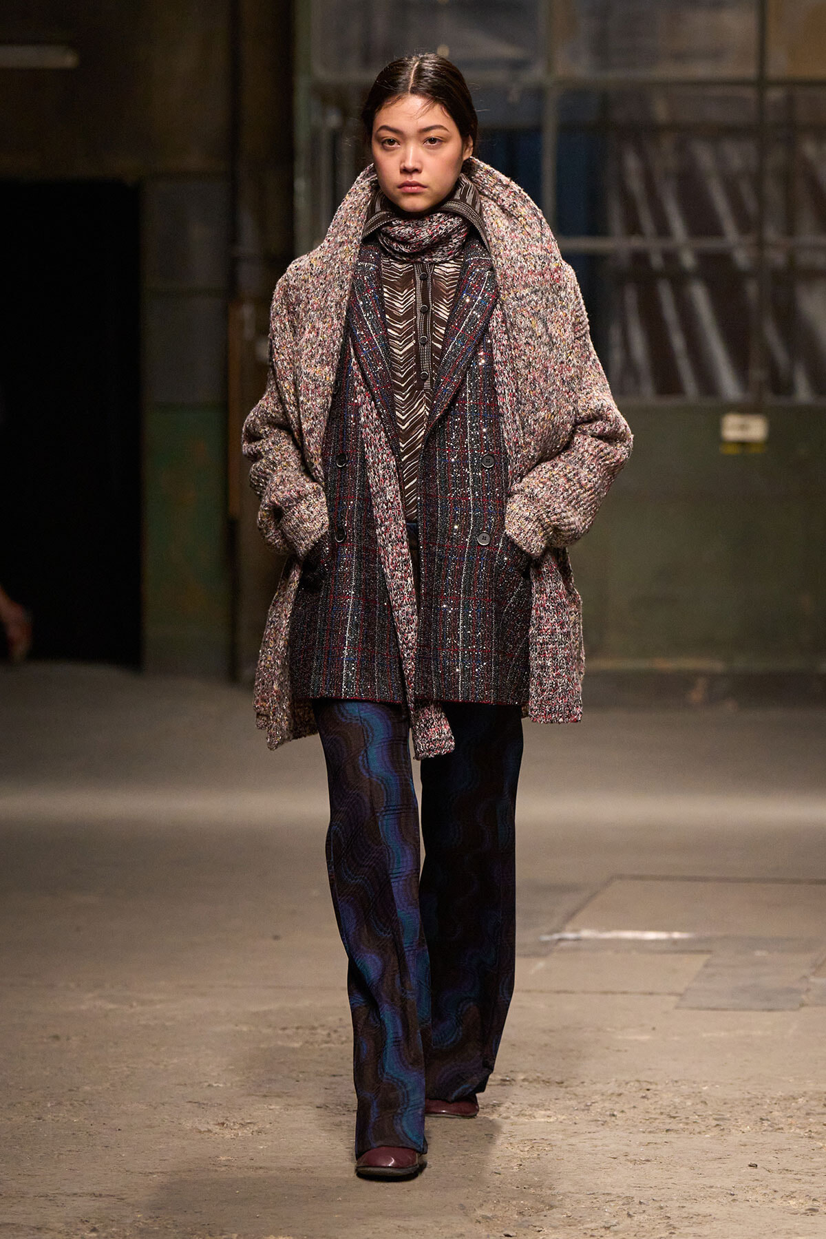 Model walking a runway in an oversized textured coat over a plaid blazer and scarf, with patterned wide-leg pants.