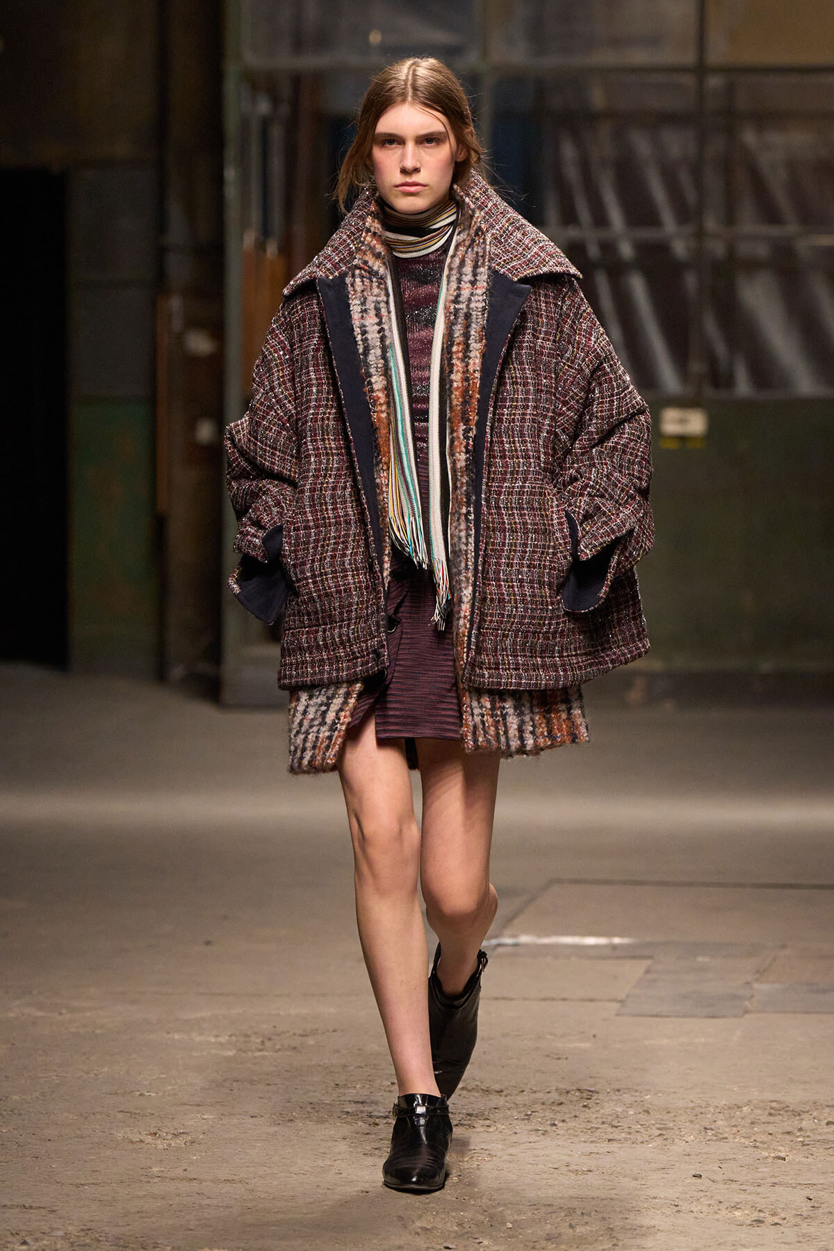 Model walking runway in an oversized plaid coat with a multicolored scarf and dark ankle boots in an urban backstage setting.