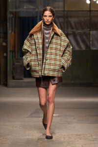 Model walks a runway in an oversized plaid coat with a brown suede collar, layered over a striped turtleneck and burgundy mini skirt, finishing with glittery flats