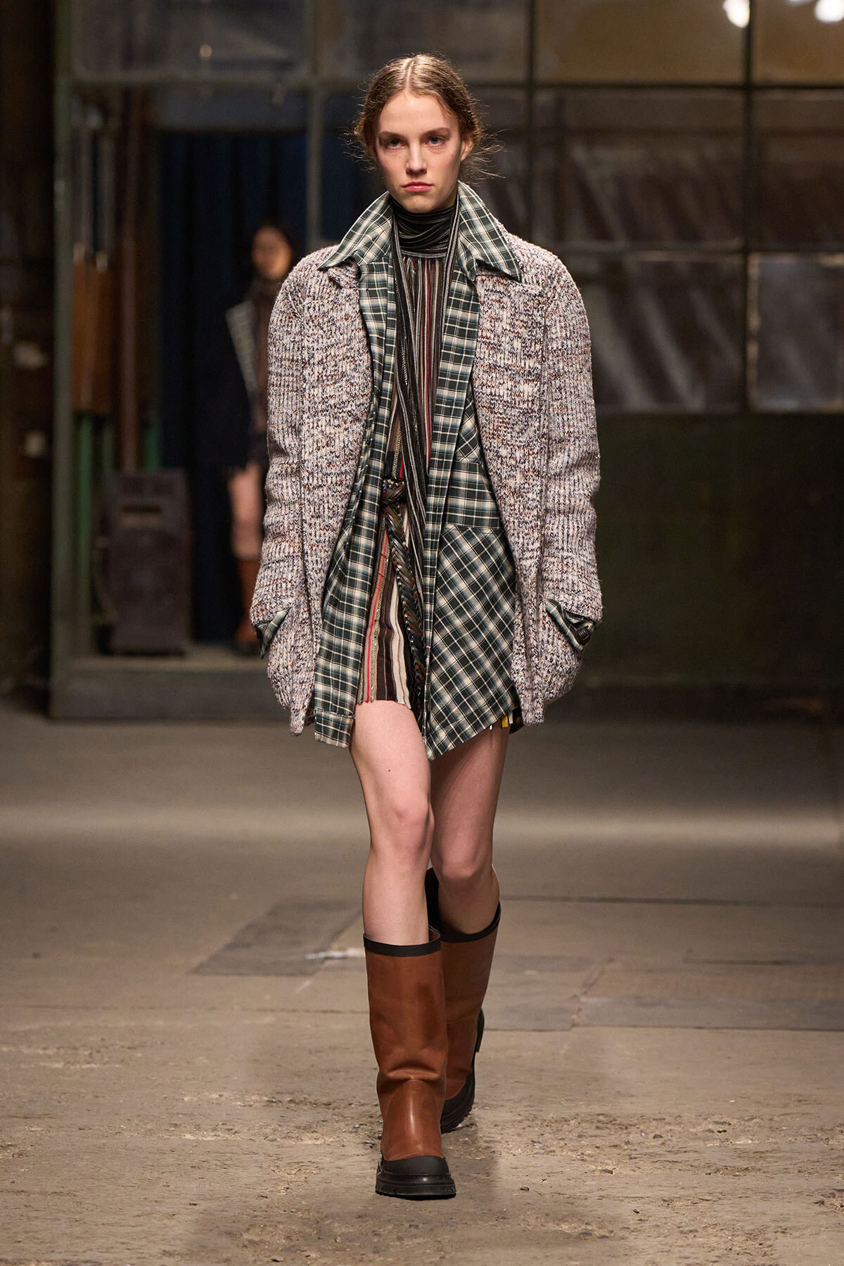 Model walking a runway in a chunky gray tweed coat over layered plaid tops and a striped dress, paired with brown knee-high boots.