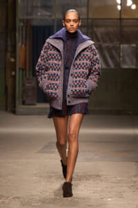 Model walking a runway in an oversized multicolor plaid knit jacket over a dark turtleneck and a fringed mini skirt, brown ankle boots.