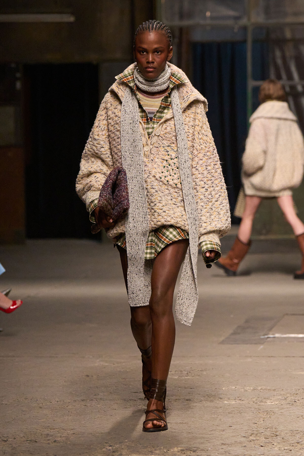 Model walking a fashion runway in a cream chunky cardigan over a plaid shirt, with a long gray scarf and brown sandals.