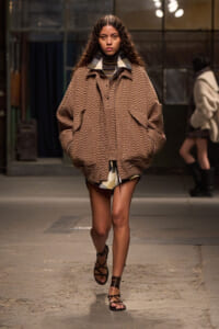 Model walks a runway in a bulky brown zigzag coat over a striped turtleneck and shorts, pairing with strappy sandals.