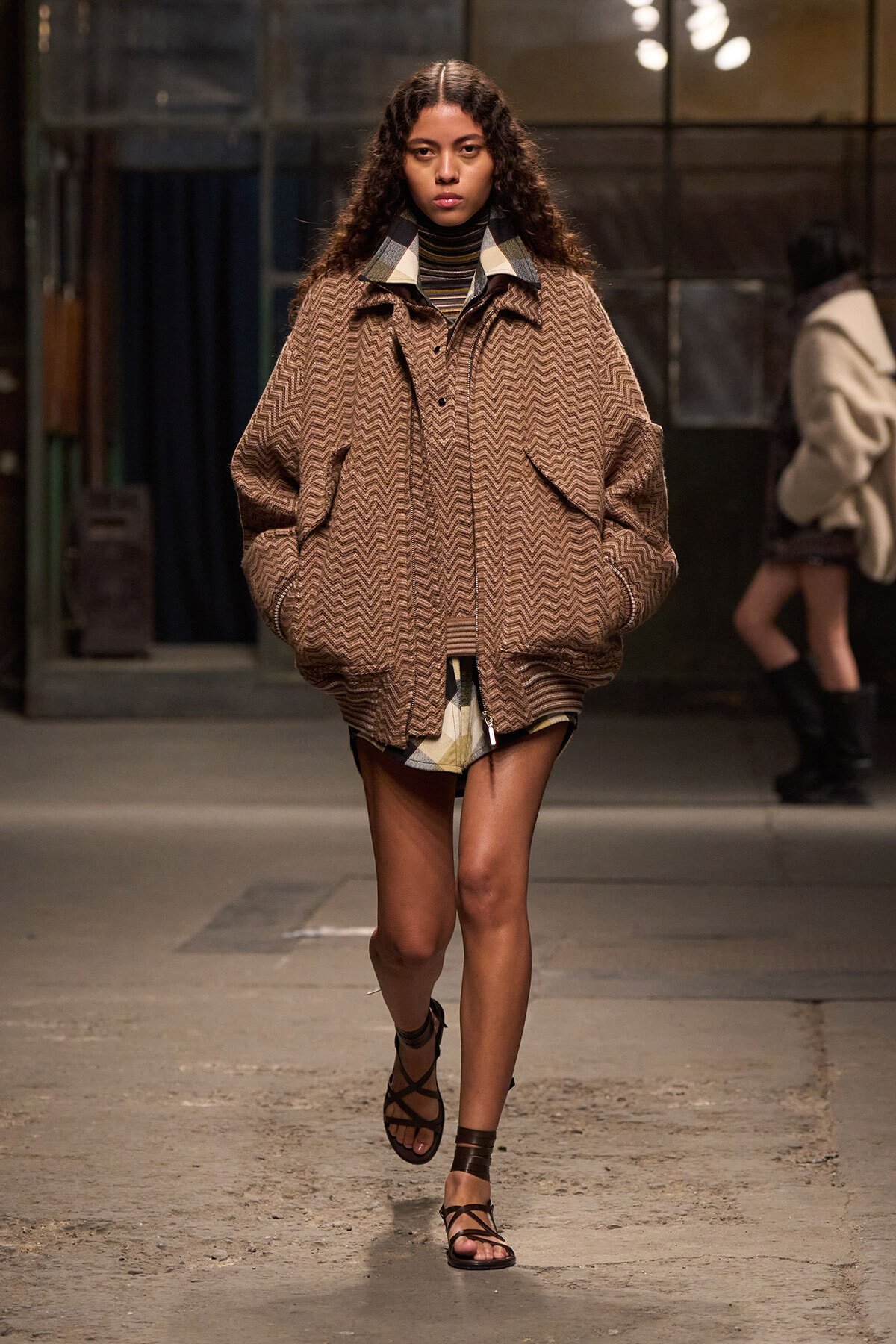 Model walks a runway in a bulky brown zigzag coat over a striped turtleneck and shorts, pairing with strappy sandals.
