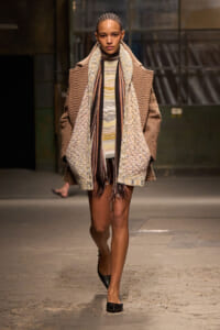 Model walking a runway in oversized, textured brown coat layered over a patterned knit sweater and scarf.