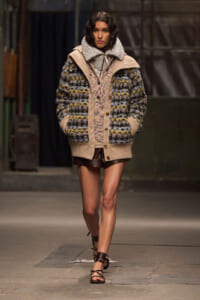 Model on a runway wearing an oversized color-block knit cardigan with a shearling collar, paired with brown leather shorts and strappy sandals.