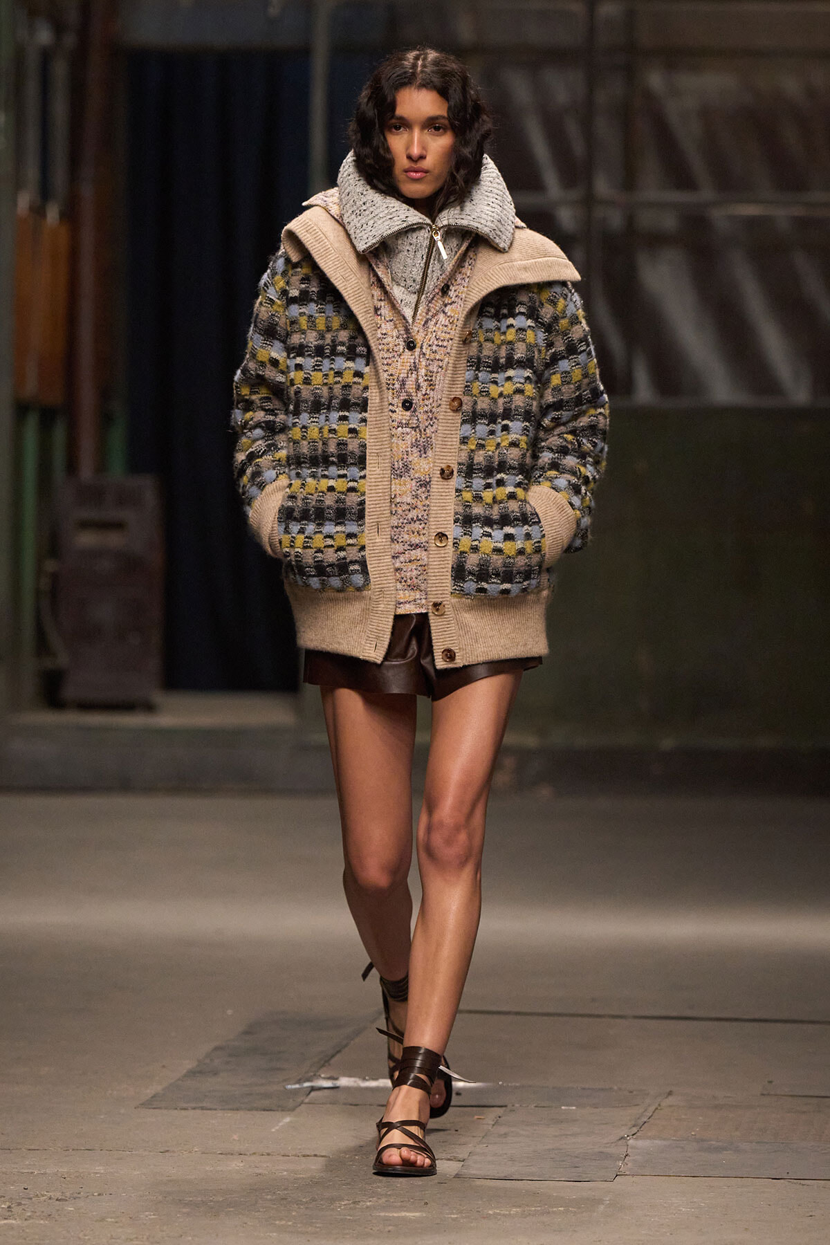 Model on a runway wearing an oversized color-block knit cardigan with a shearling collar, paired with brown leather shorts and strappy sandals.