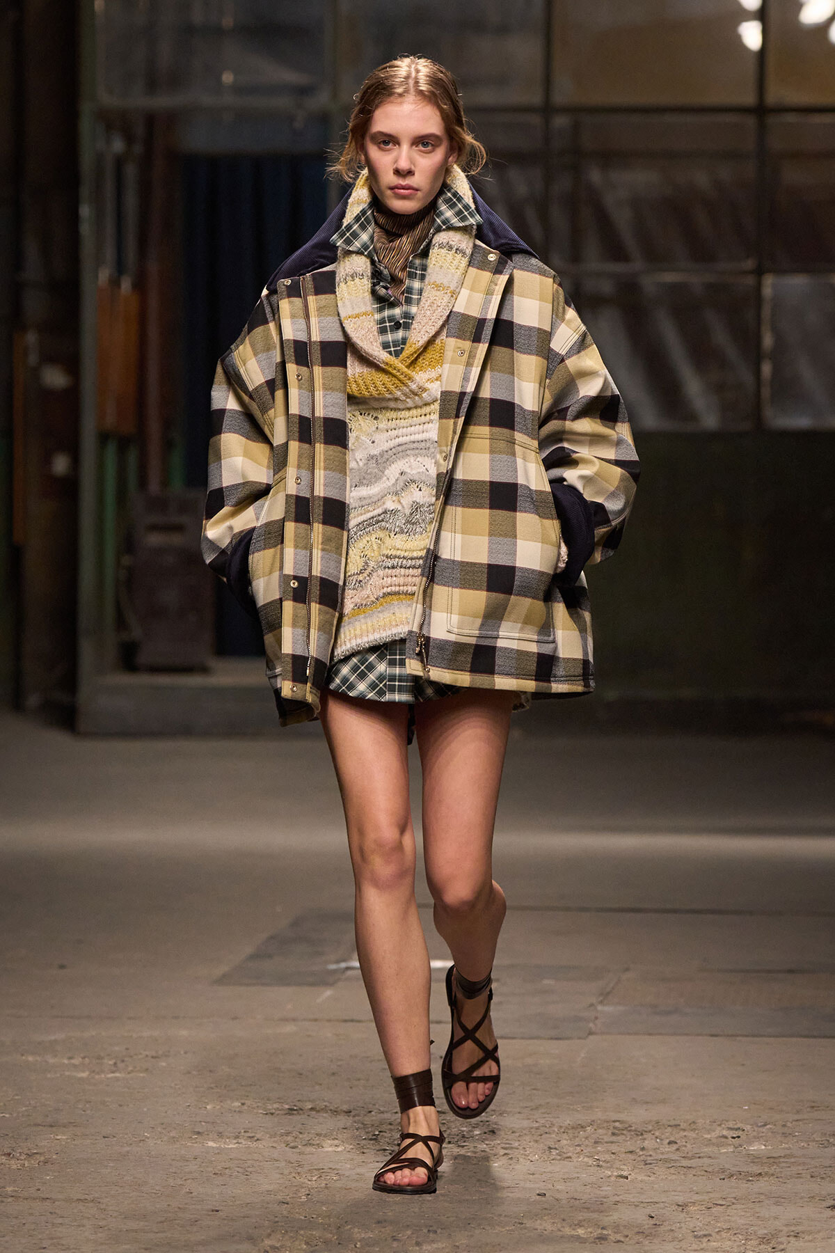 Model walks a runway in an oversized beige, plaid coat over a chunky knit sweater and plaid dress, wearing strappy brown sandals.