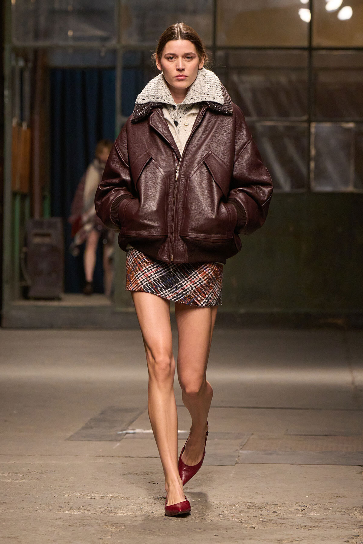 Model walking a fashion show runway in a dark brown shearling-collar leather bomber, plaid mini skirt, and burgundy flats; hands in pockets.