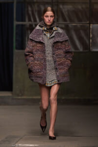 Model walking a fashion runway in an oversized textured coat layered over a plaid shirt and gray sweater, mid-stride.