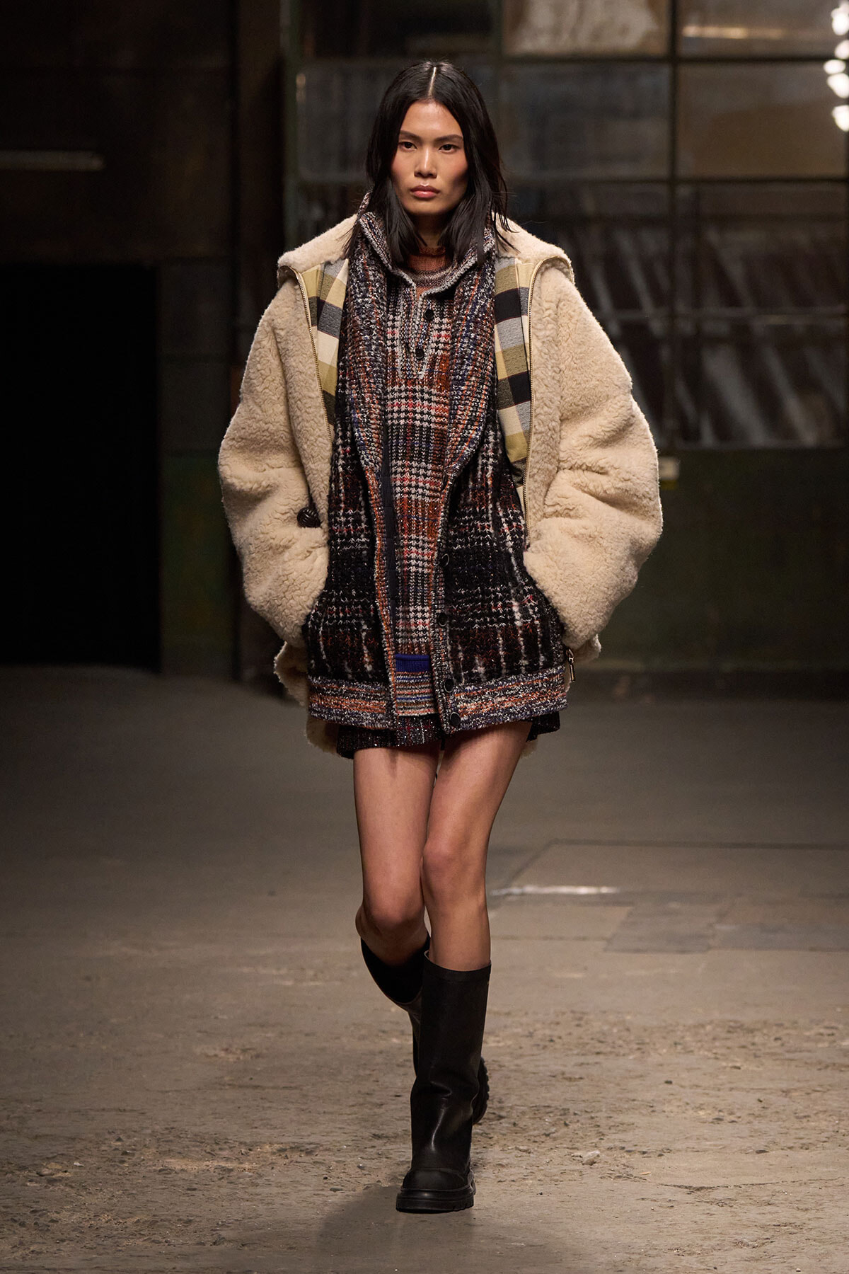 Model walks a runway in an oversized cream shearling coat over a plaid knit outfit with black knee-high boots.