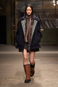 Model walking a runway in an oversized navy coat with plaid inset panels, a patterned scarf, and brown knee-high boots.