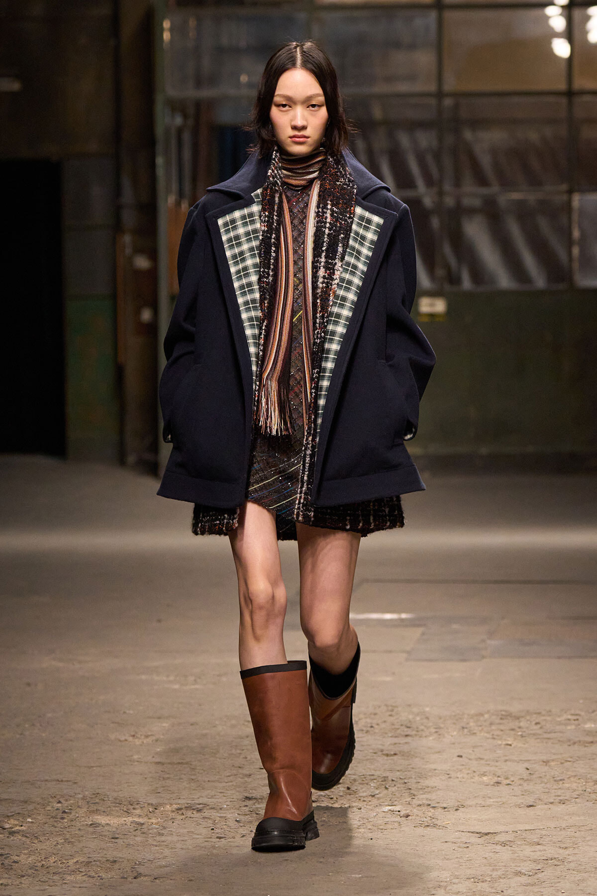 Model walking a runway in an oversized navy coat with plaid inset panels, a patterned scarf, and brown knee-high boots.