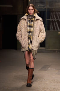 Model walking a runway in a chunky beige knit coat over a tan-and-black plaid shirt dress, paired with knee-high brown boots.