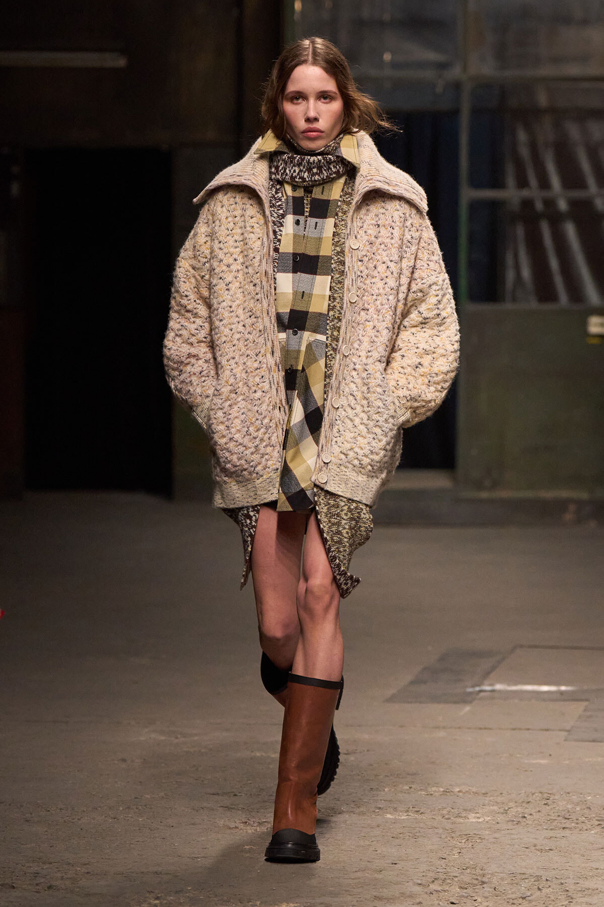 Model walking a runway in a chunky beige knit coat over a tan-and-black plaid shirt dress, paired with knee-high brown boots.