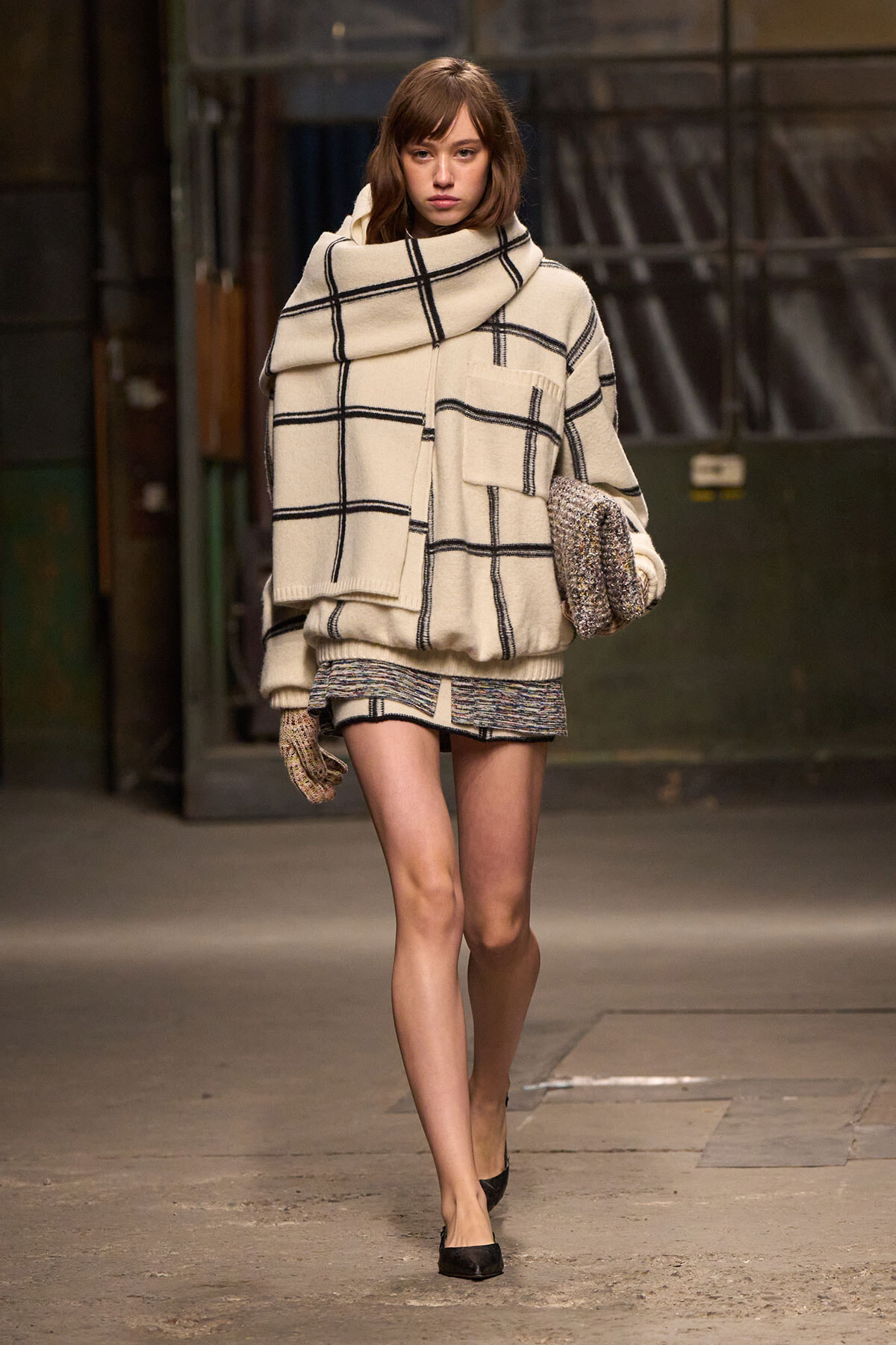 Model walking in a cream plaid oversized coat with a large scarf and gloves, carrying a textured clutch.