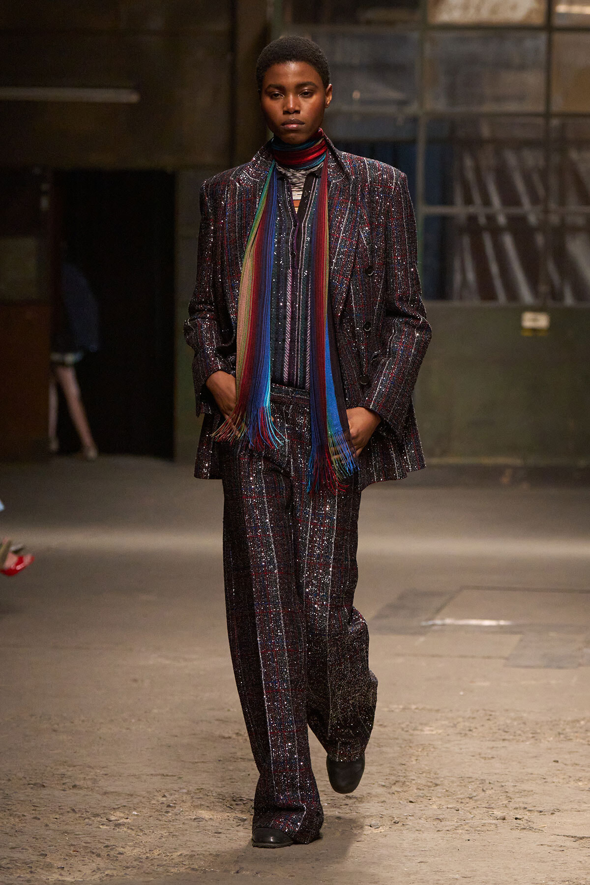 Model walks a runway in a glittery dark pinstripe suit paired with a long rainbow scarf, hands in pockets.