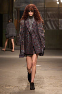 Model walking a runway in a glittery plaid oversized coat, gray turtleneck, black sunglasses, and black ankle boots.