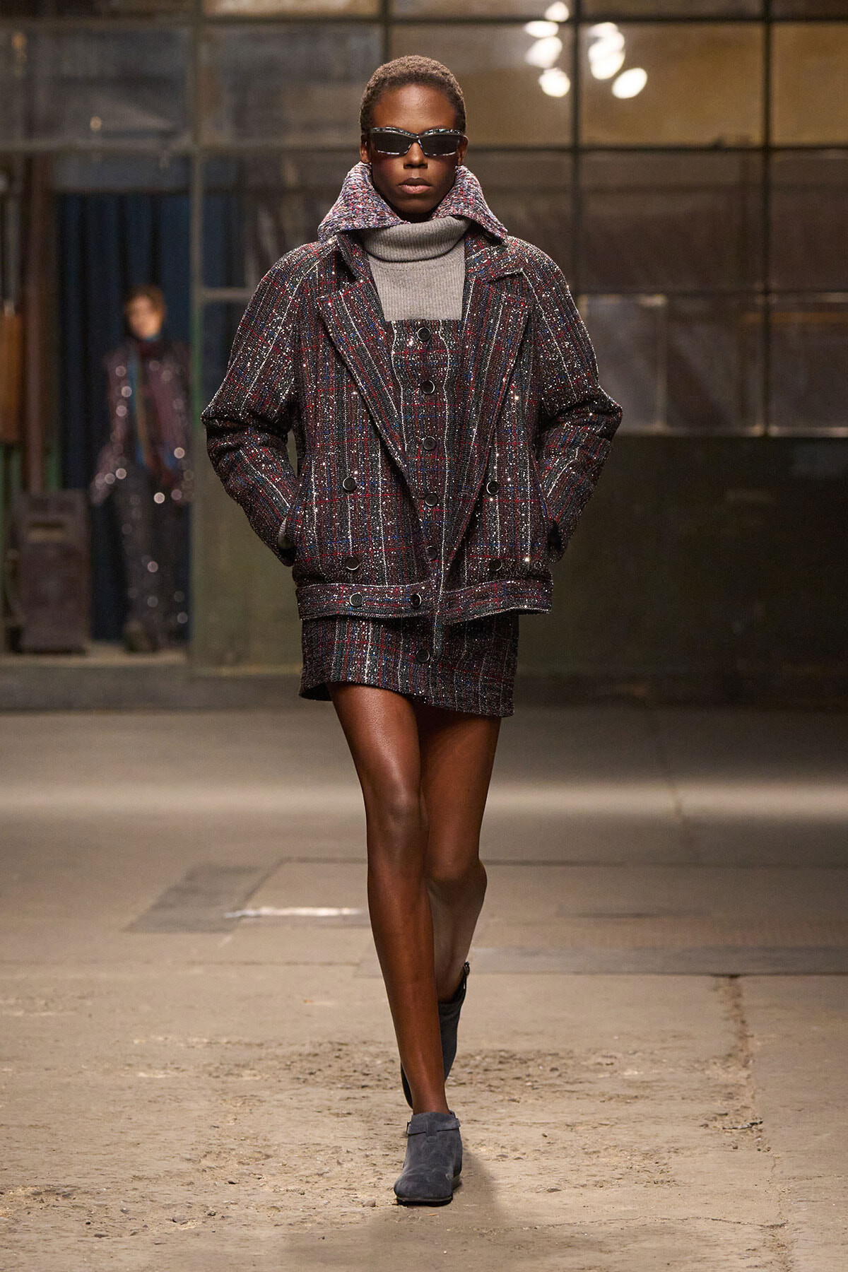 Model walks runway in a dark, plaid blazer and matching mini skirt, gray turtleneck, and sunglasses; urban backstage setting.