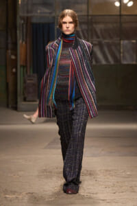 Model on a runway wearing a bold multicolor striped coat, knit top, and glittery dark pants in an industrial venue