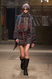 Runway model in a multicolor plaid knit coat and matching accessories, wearing a beanie, scarf, sunglasses, and black knee-high boots.