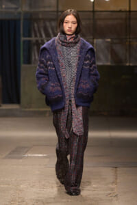 Model on a runway wearing an oversized navy cardigan over a patterned knit outfit and plaid trousers.