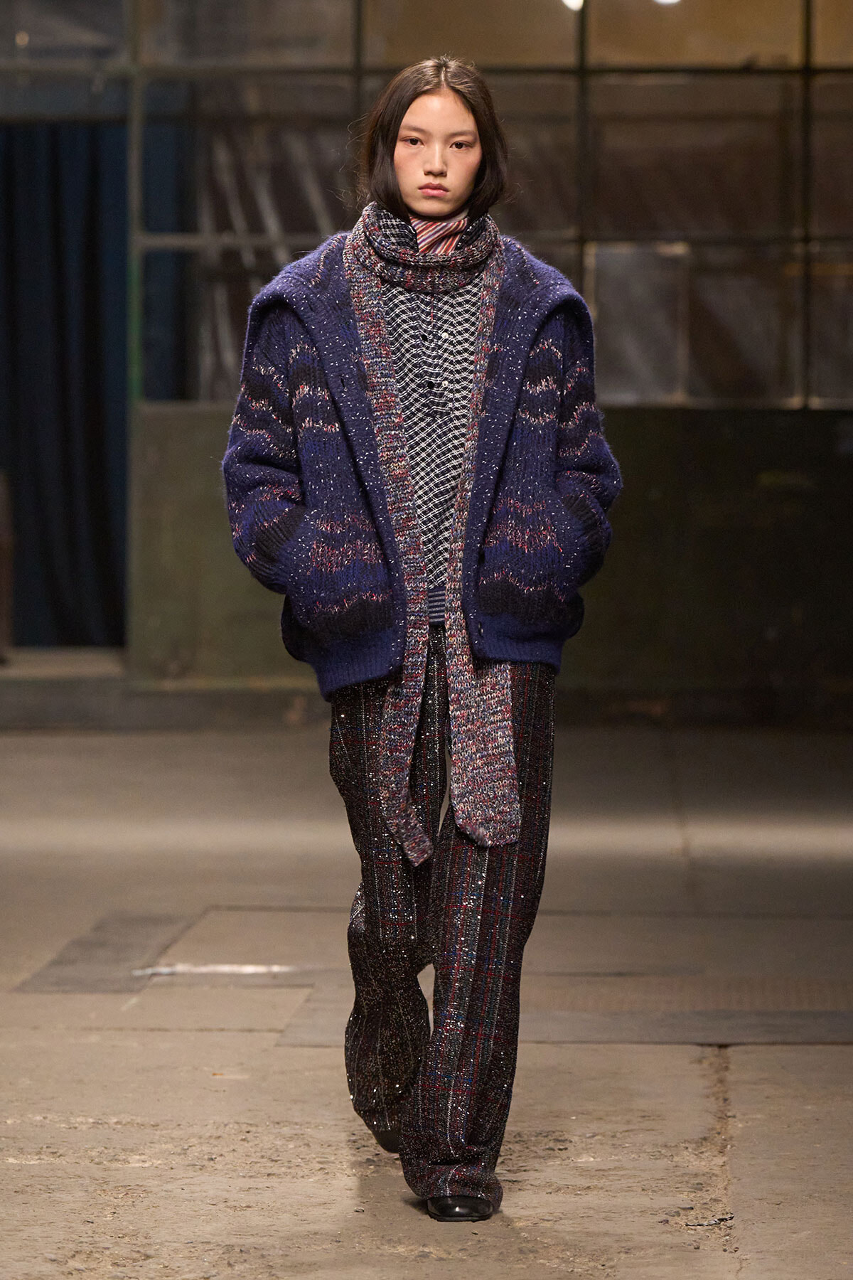 Model on a runway wearing an oversized navy cardigan over a patterned knit outfit and plaid trousers.