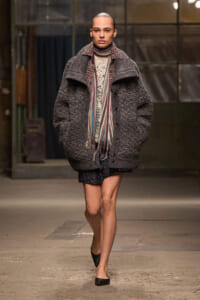 Female model on a runway wears an oversized dark gray textured coat with a multicolor striped scarf, cream knit sweater, glittery shorts, and black flats.