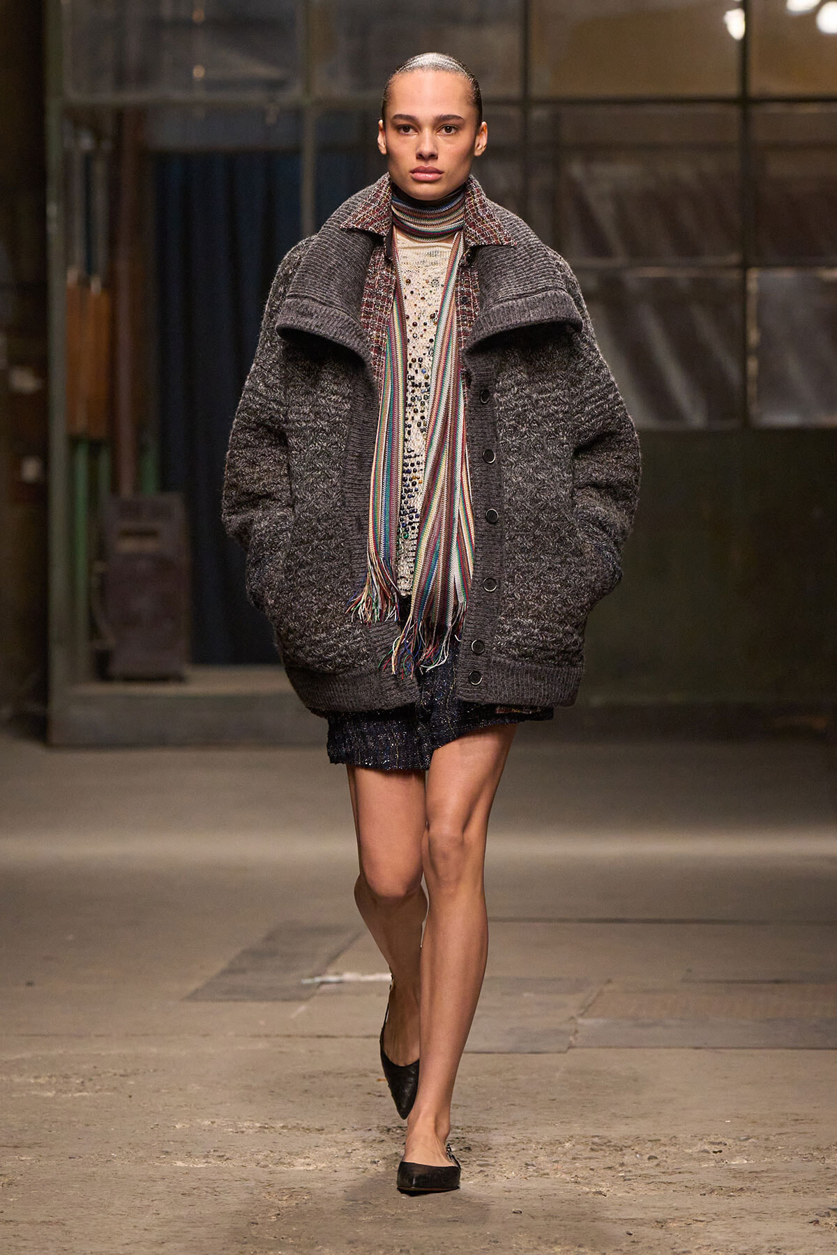 Female model on a runway wears an oversized dark gray textured coat with a multicolor striped scarf, cream knit sweater, glittery shorts, and black flats.