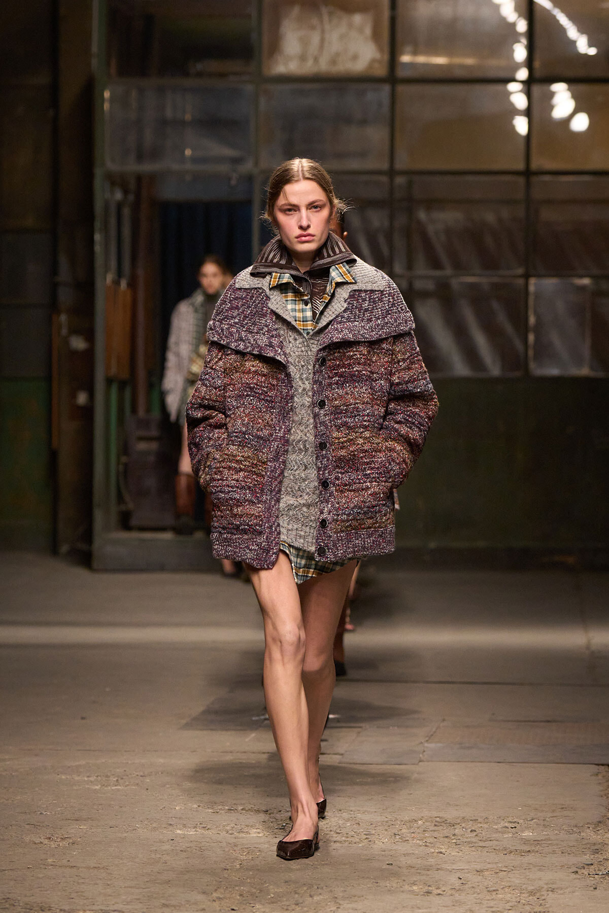 Model walking a runway in an oversized maroon-grey textured coat over a plaid shirt in an industrial backstage setting.