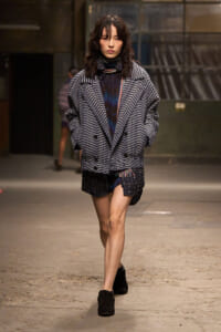 Model walking toward camera in an oversized plaid coat over a dark dress, black boots, in an industrial venue runway setting