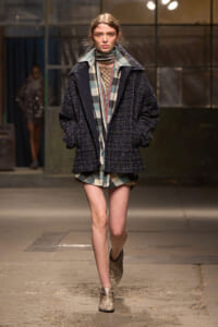 Model walks the runway in a dark, oversized textured navy coat layered over plaid pieces and ankle boots.